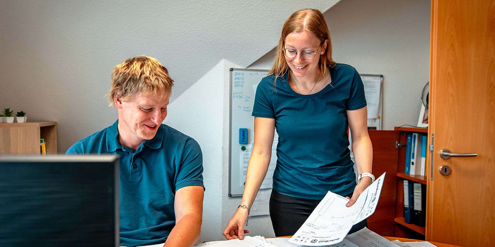 Ein Mann und eine Frau in blauen T-Shirts arbeiten zusammen an einem Tisch, umgeben von Dokumenten und Büromaterial.