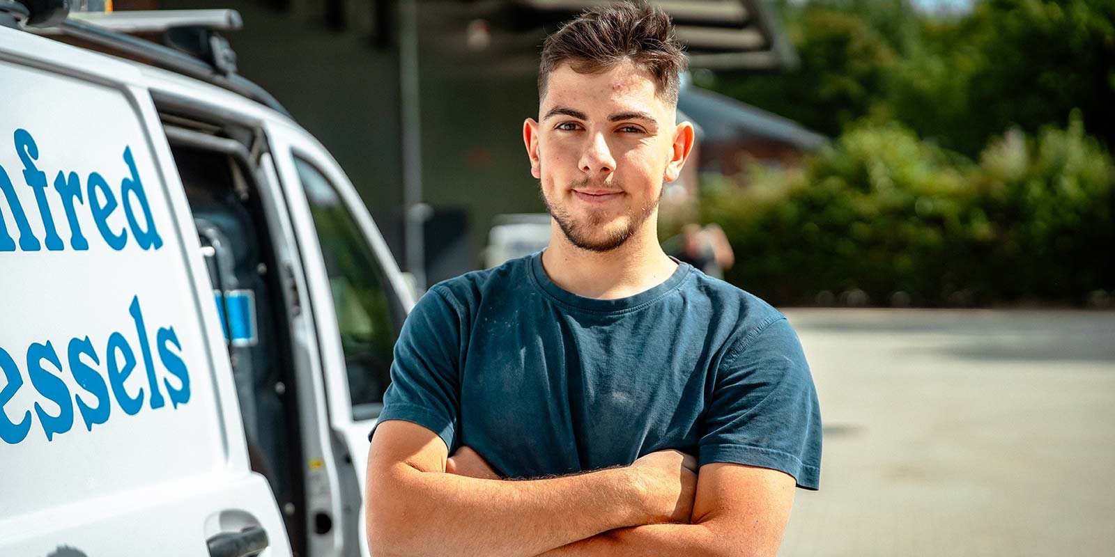 Ein junger Mann mit einem lächelnden Gesicht steht vor einem Lieferwagen, die Arme vor der Brust verschränkt.