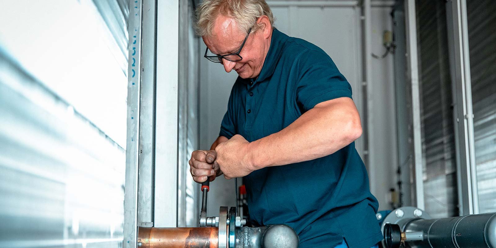 Ein Mann mit Brille arbeitet konzentriert an einem Rohr, während er einen Schraubenschlüssel in der Hand hält.
