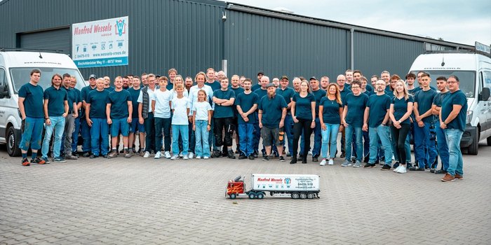 Eine große Gruppe von Mitarbeitern steht vor einem Firmengebäude, alle tragen einheitliche dunkelblaue T-Shirts und lächeln.