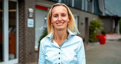 Eine lächelnde Frau mit blonden Haaren steht vor einem modernen Gebäude, das eine freundliche Atmosphäre ausstrahlt.