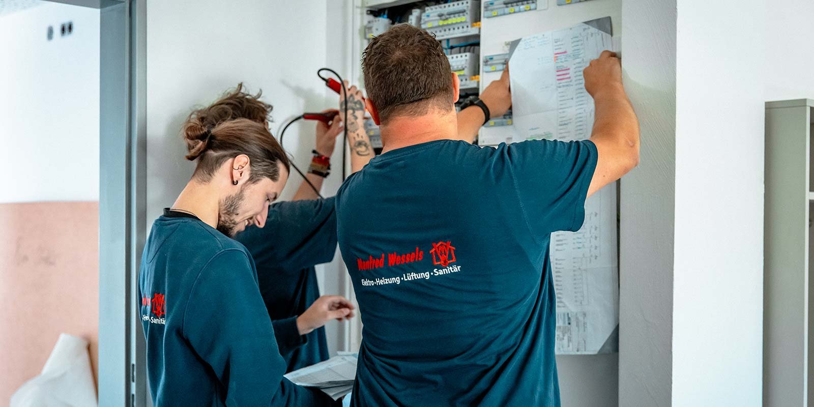 Zwei Techniker arbeiten konzentriert an einem elektrischen Schaltschrank, während sie Pläne und Werkzeuge verwenden.