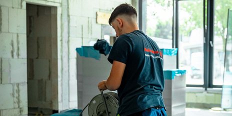 Ein junger Mann in einem schwarzen T-Shirt arbeitet konzentriert in einem hellen Raum mit großen Fenstern.
