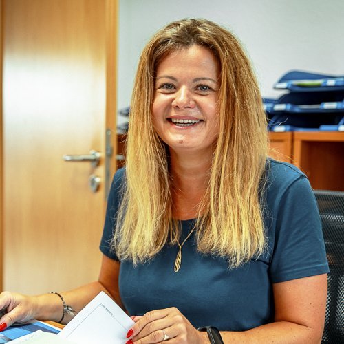 Eine lächelnde Frau mit langen, blonden Haaren sitzt an einem Schreibtisch und hält Dokumente in der Hand.
