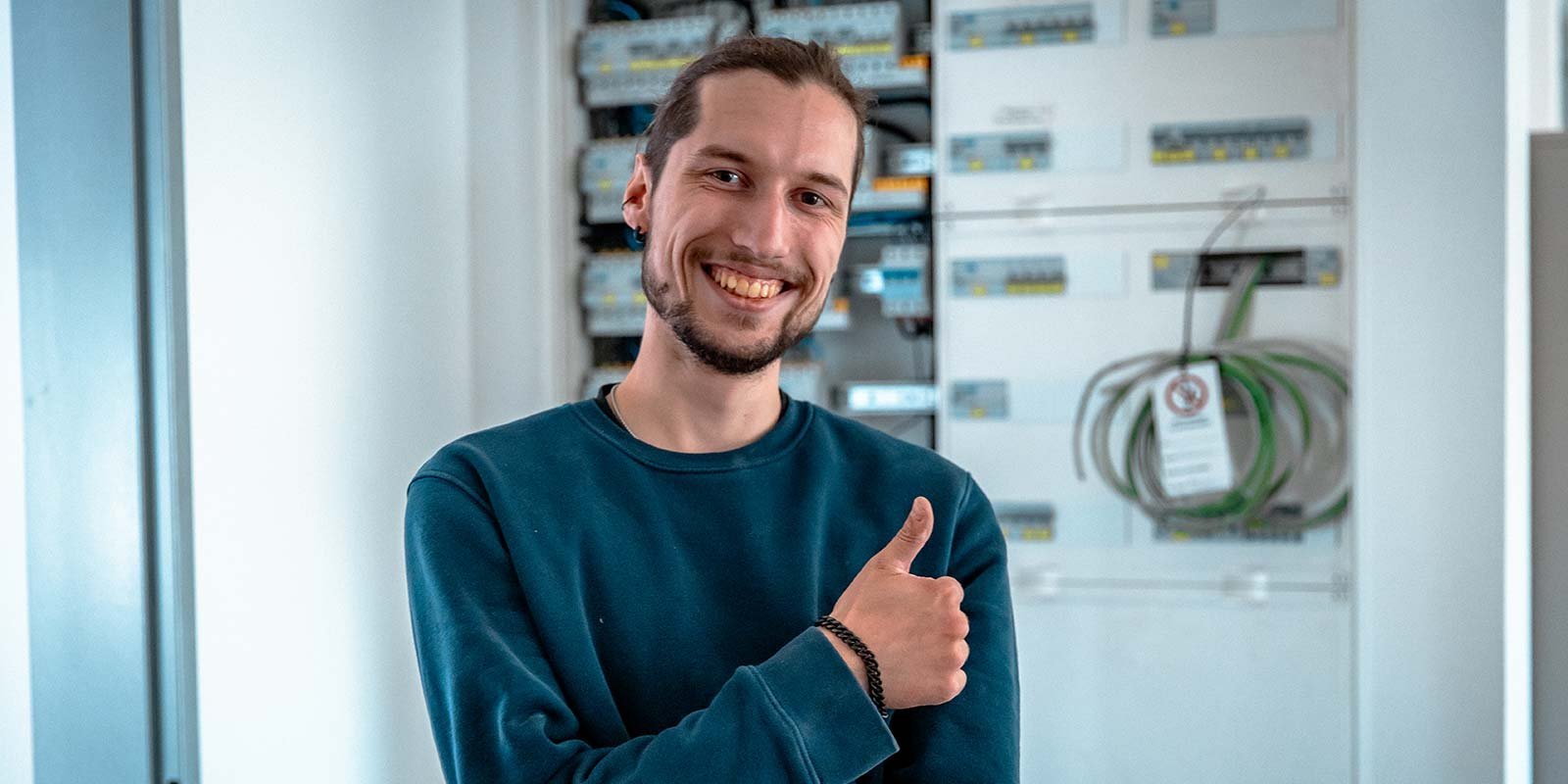 Ein junger Mann mit einem breiten Lächeln steht vor einem elektrischen Schaltschrank und zeigt Daumen hoch.