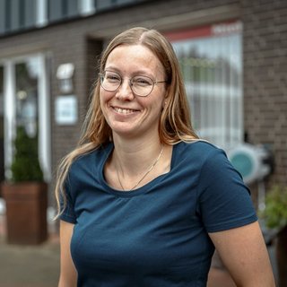 Lächelnde Frau vor einem modernen Gebäude Eine lächelnde Frau mit Brille steht vor einem modernen Gebäude, umgeben von Pflanzen und einem freundlichen Ambiente.