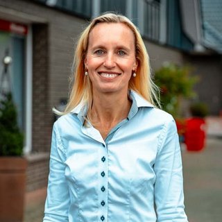 Lächelnde Frau in blauer Bluse vor modernem Gebäude Eine blonde Frau mit langen Haaren lächelt freundlich vor einem modernen Gebäude, das mit Pflanzen dekoriert ist.