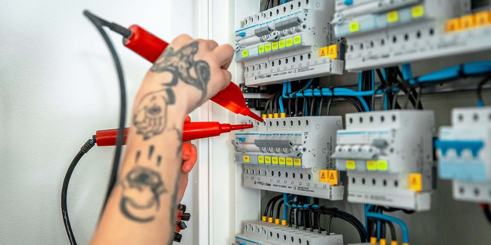 Eine Person mit einem tätowierten Arm verwendet rote Prüfspitzen, um elektrische Schaltkästen zu überprüfen.