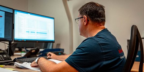 Ein Mann sitzt an einem Schreibtisch und arbeitet konzentriert an zwei Computermonitoren mit technischen Daten.