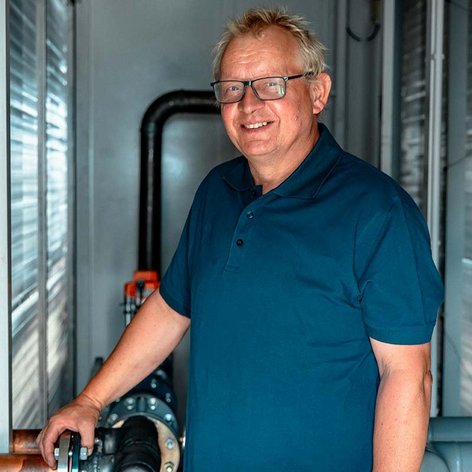 Ein lächelnder Mann in einem blauen Poloshirt steht neben einer Rohrleitung in einem industriellen Umfeld.