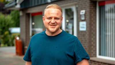 Ein lächelnder Mann mit blonden Haaren steht vor einem Gebäude mit großen Fenstern und einem modernen Design.