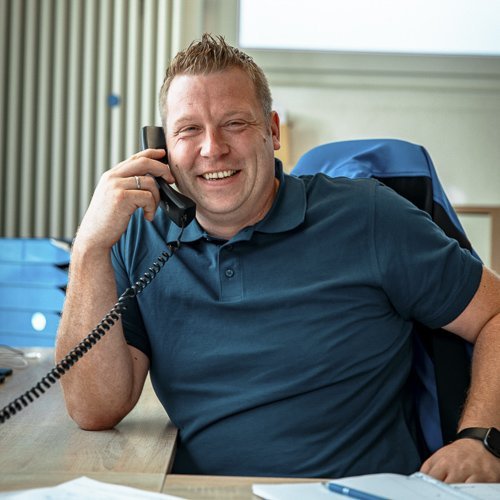 Ein lächelnder Mann spricht am Telefon, während er an einem Schreibtisch mit Aktenordnern sitzt.