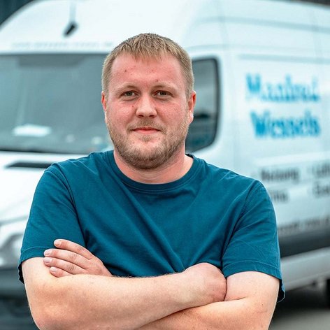 Ein Mann mit kurzem Haar und Bart steht mit verschränkten Armen vor einem Lieferwagen, der mit einem Logo versehen ist.