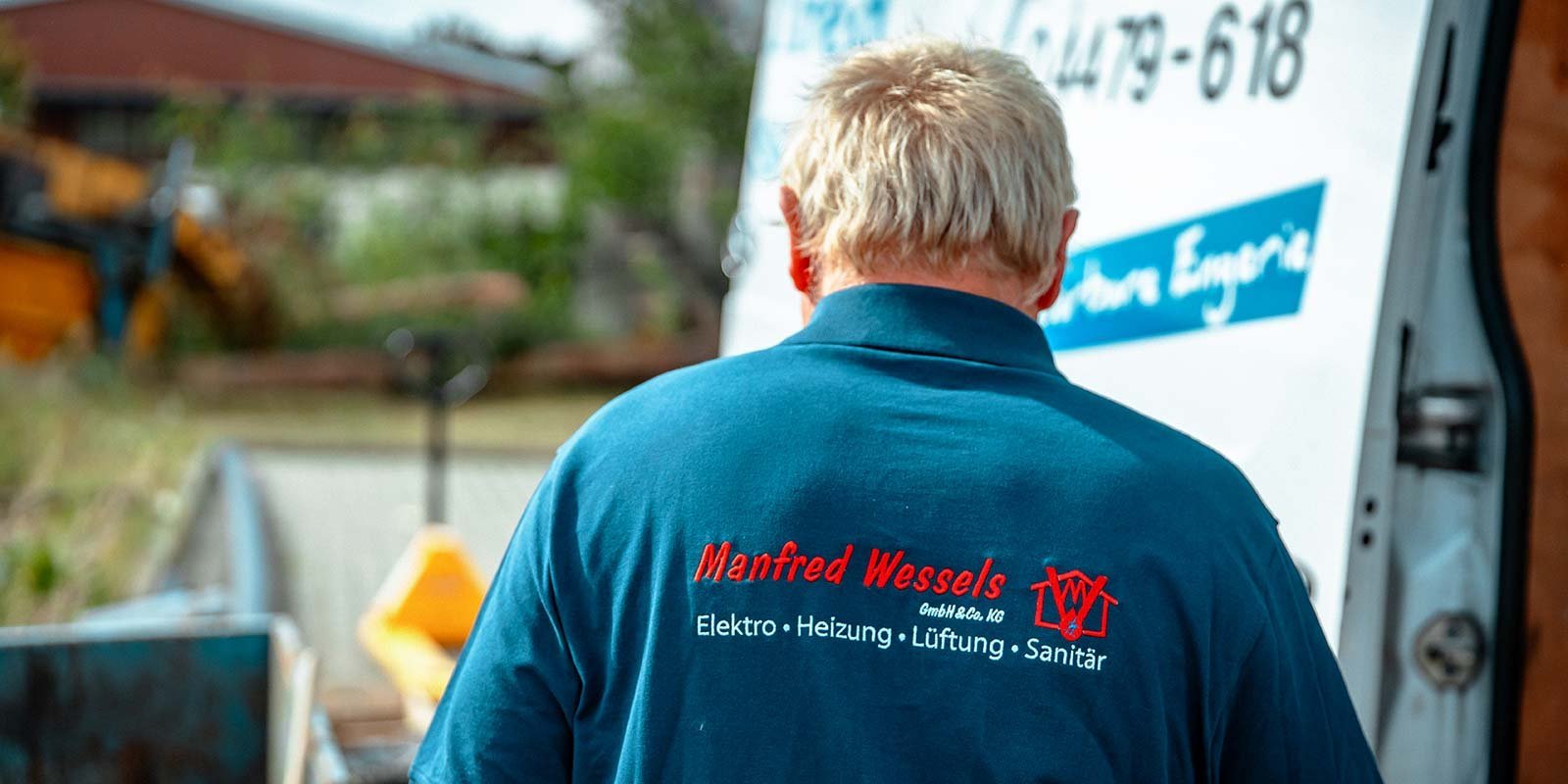 Ein Mann mit blonden Haaren trägt ein blaues Shirt mit dem Namen 'Manfred Wessels' und steht vor einem Lieferwagen.