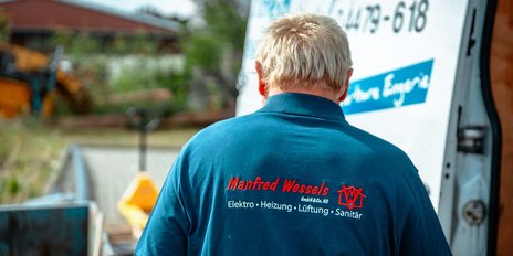 Ein Mann mit blonden Haaren trägt ein blaues Shirt mit dem Namen 'Manfred Wessels' und steht vor einem Lieferwagen.