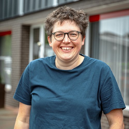 Eine lächelnde Frau mit kurzen, lockigen Haaren trägt ein blaues T-Shirt und steht vor einem Gebäude mit großen Fenstern.