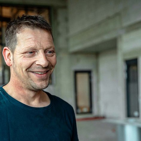 Ein lächelnder Mann mit kurzen Haaren steht in einem modernen, hellen Raum mit großen Fenstern im Hintergrund.