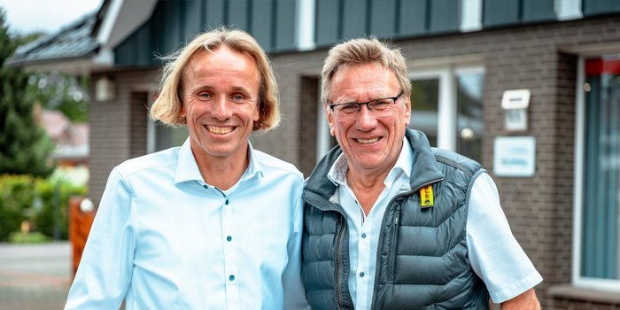 Zwei Männer stehen lächelnd vor einem modernen Gebäude, beide tragen helle Hemden und wirken freundlich und entspannt.