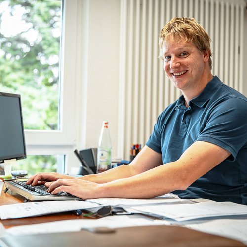 Ein lächelnder Mann sitzt an einem Schreibtisch, während er an einem Computer arbeitet, umgeben von Unterlagen und Büromaterial.