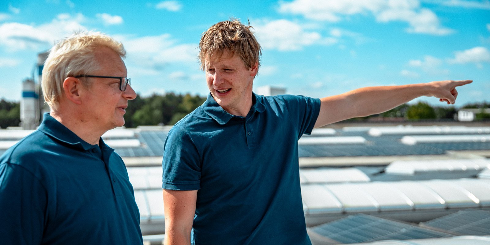 Zwei Männer in blauen Poloshirts stehen auf einem Dach und diskutieren, während sie auf die Umgebung zeigen.