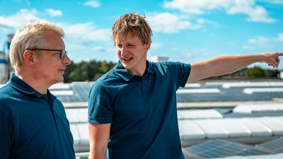 Zwei Männer in blauen Poloshirts stehen auf einem Dach und diskutieren, während sie auf die Umgebung zeigen.