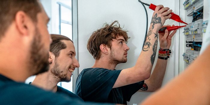 Drei junge Männer arbeiten konzentriert an einem elektrischen Schaltschrank, während sie Messgeräte in der Hand halten.