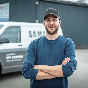 Ein junger Mann mit einem lächelnden Gesicht steht mit verschränkten Armen vor einem Lieferwagen in einer urbanen Umgebung.