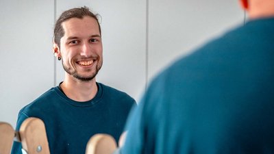 Ein junger Mann mit einem freundlichen Lächeln steht vor einem Spiegel und blickt in die Kamera.