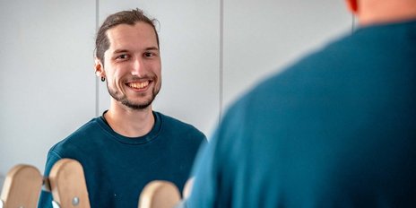 Ein junger Mann mit einem freundlichen Lächeln steht vor einem Spiegel und blickt in die Kamera.
