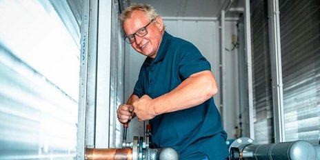 Ein Handwerker mit Brille lächelt, während er an einer Rohrverbindung mit einem Schraubenschlüssel arbeitet.