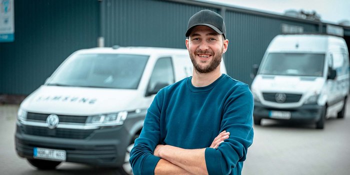 Ein lächelnder Mann mit Bart steht mit verschränkten Armen vor zwei Lieferwagen in einem industriellen Umfeld.