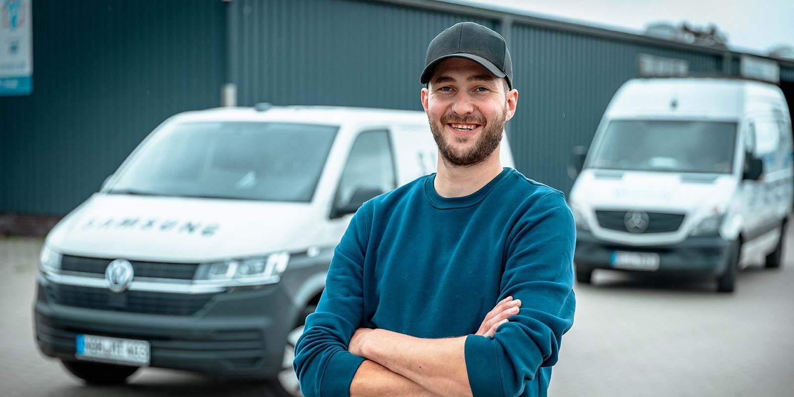 Ein lächelnder Mann mit Bart steht mit verschränkten Armen vor zwei Lieferwagen in einem industriellen Umfeld.