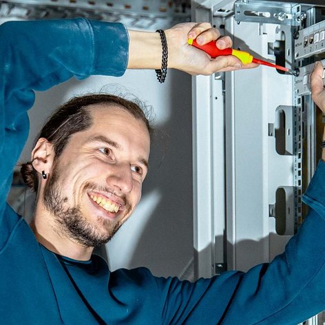 Ein Techniker arbeitet konzentriert an einem Kabelschrank und lächelt, während er mit einem Schraubendreher Kabel anschließt.