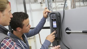 Zwei Männer konzentrieren sich auf ein technisches Gerät, während einer von ihnen einen Knopf drückt und Anweisungen gibt.