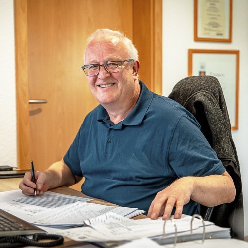 Ein lächelnder Mann mit Brille sitzt an einem Schreibtisch und arbeitet an Dokumenten, umgeben von einem hellen Raum.