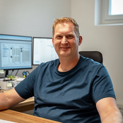 Ein lächelnder Mann sitzt an einem Schreibtisch, umgeben von zwei Computermonitoren in einem hellen Büro.