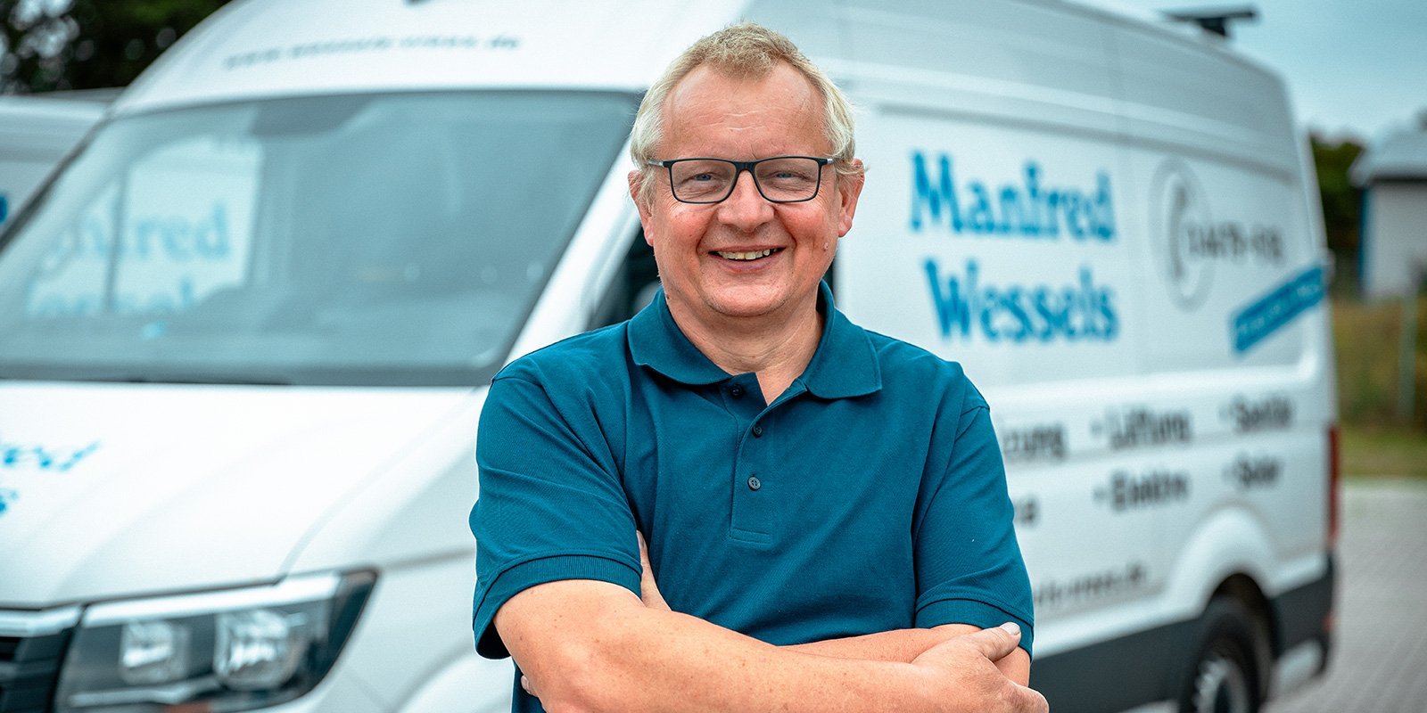 Ein lächelnder Mann mit Brille steht vor einem Lieferwagen, der mit dem Namen 'Manfred Wessels' beschriftet ist.