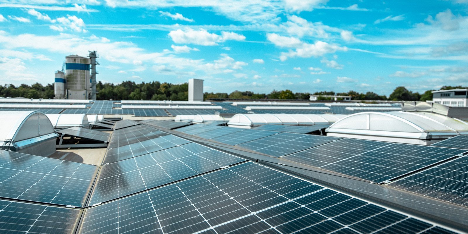 Dach mit zahlreichen Solarpanelen, die in der Sonne glänzen, während im Hintergrund ein Industriegebäude sichtbar ist.