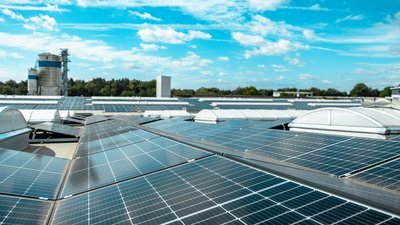 Dach mit zahlreichen Solarpanelen, die in der Sonne glänzen, während im Hintergrund ein Industriegebäude sichtbar ist.