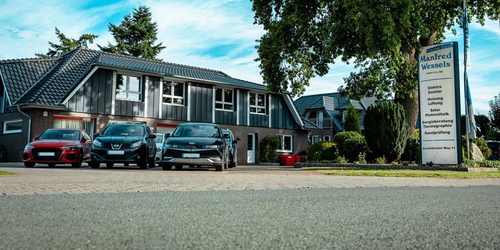 Drei Autos parken vor einem modernen Gebäude mit einem großen Schild, das die Dienstleistungen von Manfred Wessels anzeigt.