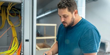Ein Mann mit kurzem, lockigem Haar arbeitet konzentriert an einem Kabelschrank, umgeben von bunten Kabeln.