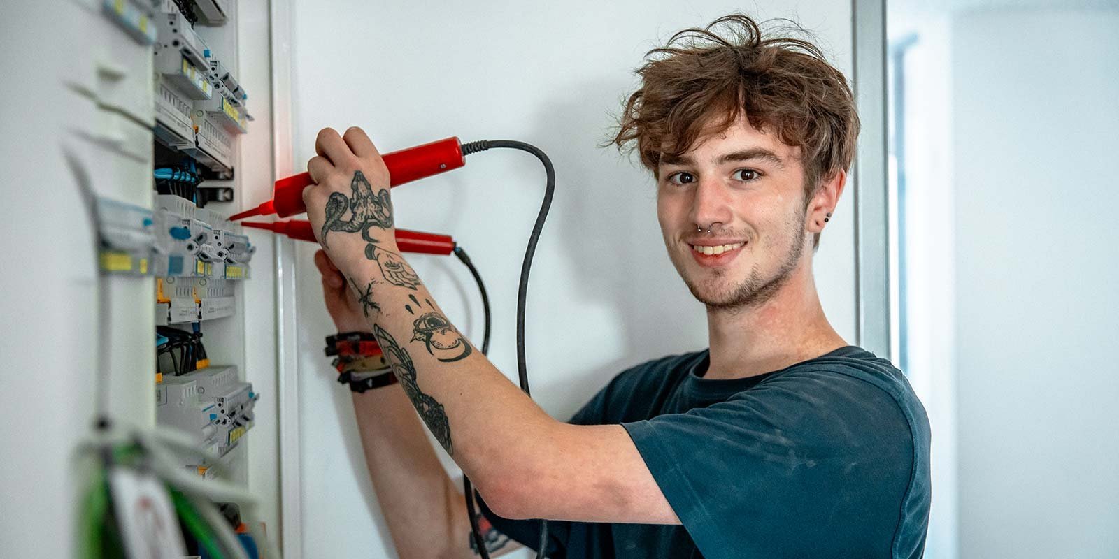 Ein junger Mann mit Tattoos lächelt, während er mit einem Multimeter an einem elektrischen Schaltschrank arbeitet.