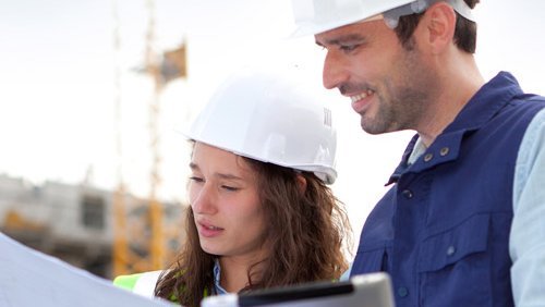 Eine Frau mit Bauhelm hält eine Zeichnung neben ihr steht ein Mann mit einem Tablet auf einer Baustelle